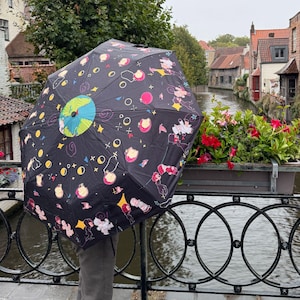 Puede incluir: Un paraguas negro con un patrón colorido de planetas, estrellas y figuras de dibujos animados. El paraguas está abierto y el mango no es visible. El fondo muestra un canal con edificios y árboles.