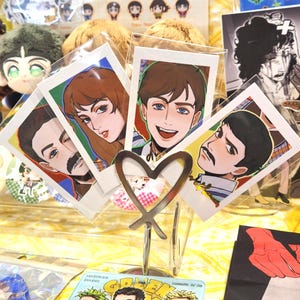 May include: Four portrait-style art prints featuring illustrations of people, displayed in a clear acrylic holder with a heart-shaped design. The prints have colorful borders and are displayed on a table with other merchandise.