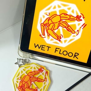 Puede incluir: Un llavero hexagonal amarillo y blanco con una figura de dibujos animados naranja, que refleja el letrero de "Precaución suelo mojado". El letrero es amarillo con texto negro y un gráfico de una persona resbalando. Un dije de estrella plateada cuelga.