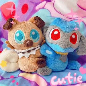 Könnte beinhalten: Zwei Plüschtiere: ein hellbrauner Welpe mit braunen Ohren und ein blau-graues Wesen mit roten Augen. Beide haben große, ausdrucksstarke Augen. Der Hintergrund ist ein farbenfrohes, gemustertes Gewebe mit dem Wort "cutie".