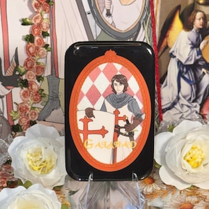 May include: Black rectangular phone case featuring a knight illustration within an orange and white checkered oval frame. The knight holds a shield with a red cross and the name "GALAHAD" is printed below. The case is displayed on a clear stand.