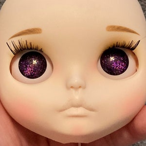 May include: A doll face with a pale complexion, detailed with brown eyebrows and long, black eyelashes. The doll's eyes are a striking purple with glitter, and the cheeks are lightly blushed. The lips are a soft, neutral color.
