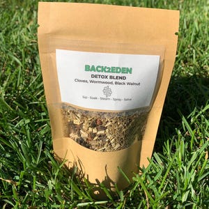 以下が含まれることがあります： 透明な窓が付いた茶色の紙袋で、ハーブデトックスブレンドが詰められています。「BACK2EDEN DETOX BLEND」と書かれたラベルには、クローブ、ヨモギ、ブラックウォールナットなどの材料が記載されています。緑の草の上に置かれています。