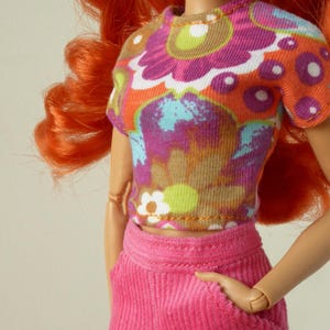 May include: A doll dressed in a floral crop top and pink corduroy trousers. The top has a pattern of purple, orange, and blue flowers. The doll has long, red hair and is posed with one hand in a pocket.