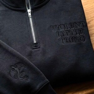 May include: Black quarter-zip sweatshirt with the embroidered text "VIOLENT LITTLE THING" on the chest. A dragon emblem is embroidered on the left sleeve. The sweatshirt has a silver zipper.