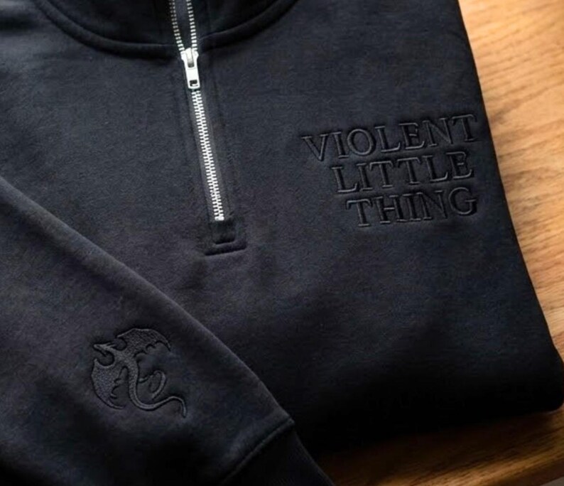 May include: Black quarter-zip sweatshirt with the embroidered text "VIOLENT LITTLE THING" on the chest. A dragon emblem is embroidered on the left sleeve. The sweatshirt has a silver zipper.