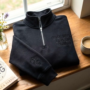 May include: Black quarter-zip sweatshirt with the text "VIOLENT LITTLE THING" embroidered on the chest. The left sleeve features an embroidered dragon design. The sweatshirt is folded on a wooden surface near a window.