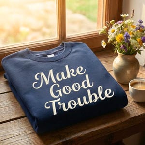 May include: A navy blue sweatshirt with the embroidered phrase "Make Good Trouble" in white script. The folded sweatshirt is on a wooden surface, next to a vase of flowers and a cup of coffee.