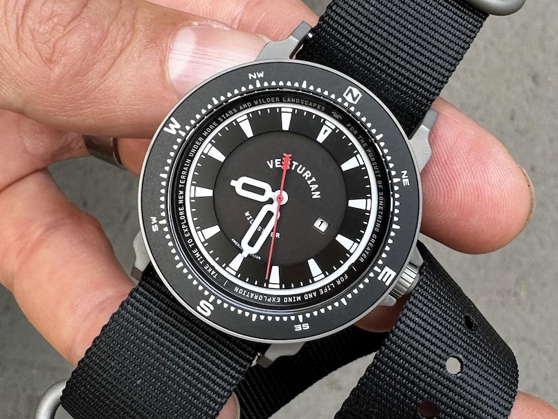 May include: Black wristwatch with a black dial and a black nylon strap. The watch face features white numerals, hands, and text, including "Veturian." The watch has a compass-style bezel and a red second hand. The watch is designed for exploration.