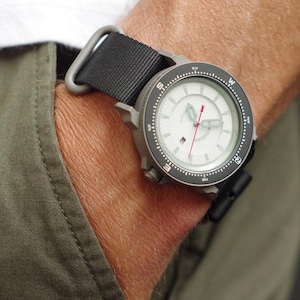 May include: A wristwatch with a white face, black bezel, and black nylon strap. The watch has a compass design and a red second hand. The watch is on a wrist, with a green pocket visible.