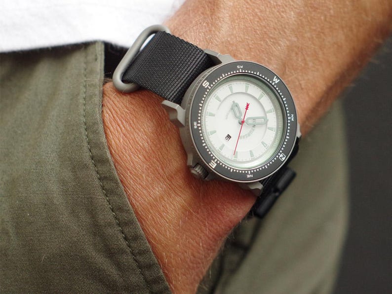 May include: A wristwatch with a white face, black bezel, and black nylon strap. The watch has a compass design and a red second hand. The watch is on a wrist, with a green pocket visible.