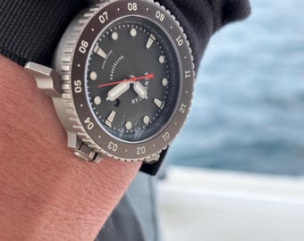 May include: A wristwatch with a dark gray dial, a rotating bezel with numbers 01 through 10, and a black strap. The watch face features the text "VENTURE" and "DAYTRIPPER" with white hour markers, a red second hand, and a date window at the six o'clock position.