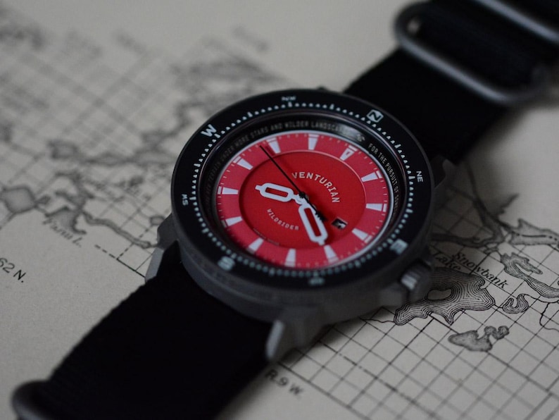 May include: A black and red VENTURIAN wristwatch with a black nylon strap. The watch face is red with white markings and hands. The watch is set against a vintage map background. The watch has the text "UNDER MORE STARS AND WILDER LANDSCAPES".