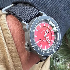 May include: A wristwatch with a red face and a black bezel is secured to a wrist with a black fabric strap. The watch has white hands and markers, and the face has the word "VILLARIAN" printed on it. The watch is on a wrist with tan fabric.