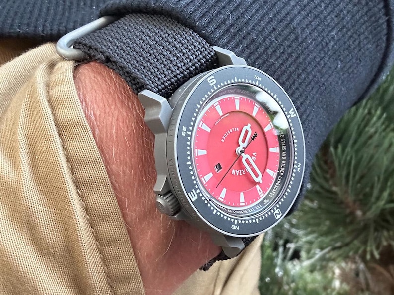 May include: A wristwatch with a red face and a black bezel is secured to a wrist with a black fabric strap. The watch has white hands and markers, and the face has the word "VILLARIAN" printed on it. The watch is on a wrist with tan fabric.