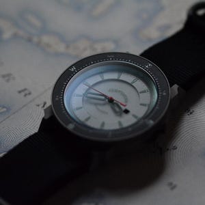 May include: A wristwatch with a black nylon strap and a silver face. The watch has a black bezel with white markings and a red second hand. The watch is resting on a map.