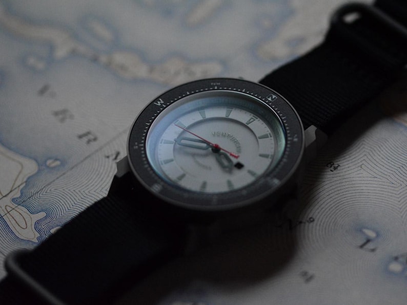 May include: A wristwatch with a black nylon strap and a silver face. The watch has a black bezel with white markings and a red second hand. The watch is resting on a map.