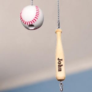 Pu&ograve; includere: Due catenelle per ventilatore a soffitto con una pallina da baseball bianca con cuciture rosse e una mazza da baseball in legno chiaro. La mazza presenta il nome John inciso in lettere nere sul lato. Entrambi pendono da catenelle metalliche.