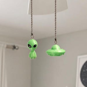 K&ouml;nnte beinhalten: Zwei Deckenventilator-Zugketten mit hellgr&uuml;nen dekorativen Ornamenten. Eine Zugkette endet in einer kleinen Alien-Figur mit gro&szlig;en schwarzen Augen, w&auml;hrend die andere Zugkette in einem gr&uuml;nen, fliegenden Untertassen-Ornament endet.