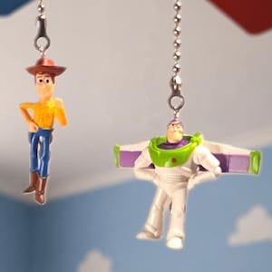 May include: Two ceiling fan pull chains featuring miniature plastic figures. One chain features a cowboy character with a brown hat, yellow shirt, and blue pants. The other chain features a space ranger character in a white, green, and purple suit.