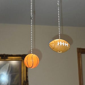 May include: Two ceiling fan pull chains featuring sports-themed ornaments. One chain ends in a round, orange basketball with black lines, while the other chain features an oval, brown football with white laces. Both are attached to metal bead chains.