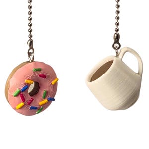 Puede incluir: Dos cadenas de tiro para ventilador de techo con dijes en miniatura. Un dije es una dona glaseada rosa con chispas de colores, y el otro es una pequeña taza de café color hueso con asa. Ambos dijes cuelgan de cadenas de bolas de metal plateado.