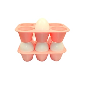 May include: A light pink silicone egg holder tray with six circular compartments arranged in two rows. The tray is stacked vertically, holding white eggs in the bottom row and one white egg in the top row, showcasing a modular design.