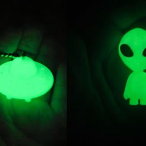 K&ouml;nnte beinhalten: Ein im Dunkeln leuchtender Schl&uuml;sselanh&auml;nger mit einem untertassenf&ouml;rmigen UFO auf der linken Seite und einer kleinen Alien-Figur auf der rechten Seite. Beide Objekte strahlen vor einem dunklen Hintergrund ein helles gr&uuml;nes Licht aus.