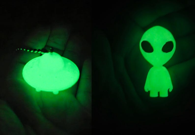 K&ouml;nnte beinhalten: Ein im Dunkeln leuchtender Schl&uuml;sselanh&auml;nger mit einem untertassenf&ouml;rmigen UFO auf der linken Seite und einer kleinen Alien-Figur auf der rechten Seite. Beide Objekte strahlen vor einem dunklen Hintergrund ein helles gr&uuml;nes Licht aus.