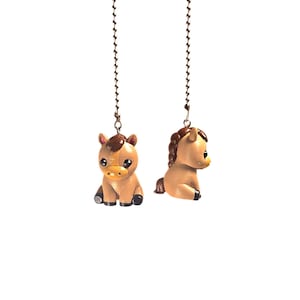May include: Two light brown horse figurines attached to silver-toned metal ball chains. Each horse has a dark brown mane, large black eyes, and dark brown hooves. One figurine faces forward, while the other is positioned in a side profile view.