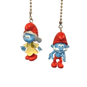 May include: Two Smurf fan pull chains. One Smurfette figure in a yellow dress and red shoes, and a Papa Smurf figure in red trousers and hat. Both figures are blue with white beards and hang from a silver chain.