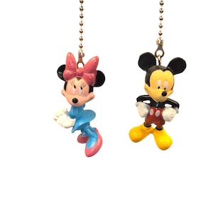 May include: Two Disney-themed fan pull chains featuring Mickey and Minnie Mouse. Mickey is in a classic pose with red shorts, yellow shoes, and white gloves. Minnie wears a blue dress, pink shoes, and a pink bow. Both are attached to a silver chain.