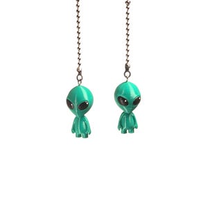 Op de afbeelding: Twee talingkleurige alien-figuurtjes bevestigd aan zilverkleurige metalen kogelkettingen. Elke alien heeft een glad, rond hoofd, grote zwarte ovale ogen en een klein lichaam met zichtbare verticale lijnen aan de voorzijde.
