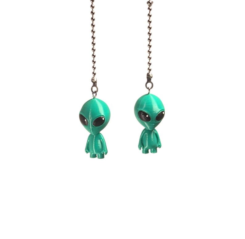Op de afbeelding: Twee talingkleurige alien-figuurtjes bevestigd aan zilverkleurige metalen kogelkettingen. Elke alien heeft een glad, rond hoofd, grote zwarte ovale ogen en een klein lichaam met zichtbare verticale lijnen aan de voorzijde.