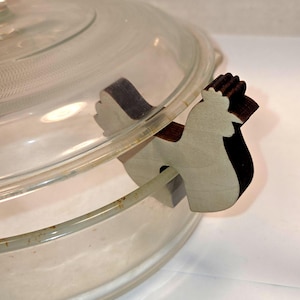 May include: A clear glass lid for a cooking vessel featuring a wooden pot minder shaped like a rooster. The pot minder is light-colored wood with dark, scorched edges and is designed to sit on the rim of the glass lid to create a small gap.