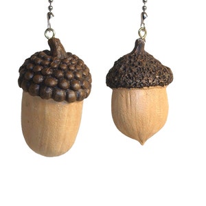 May include: Two acorn-shaped ceiling fan pulls hanging from silver-toned metal ball chains. Each acorn features a textured dark brown cap and a smooth, light tan nut body. The left acorn has a rounded cap, while the right has a rougher texture.