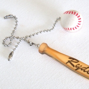May include: Baseball-themed ceiling fan pull chain featuring a baseball and a miniature wooden baseball bat with the name "Ryder" burned into the wood. The baseball is white with red stitching, and the bat is light brown.