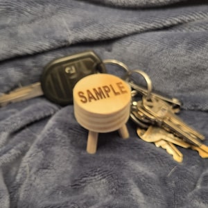May include: A wooden miniature stool with the word "SAMPLE" engraved on top. The stool is light brown and has three small legs. A set of keys and a car key are in the background, resting on a blue fabric.