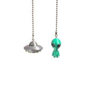 Op de afbeelding: Twee zilverkleurige metalen kogelkettingen, elk met een kleine hanger. De ene hanger is een grijze, koepelvormige UFO met kleine groene accenten langs de basis. De andere hanger is een klein, groen, mensachtig alienfiguurtje met grote zwarte ogen.