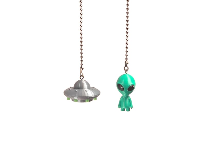 Op de afbeelding: Twee zilverkleurige metalen kogelkettingen, elk met een kleine hanger. De ene hanger is een grijze, koepelvormige UFO met kleine groene accenten langs de basis. De andere hanger is een klein, groen, mensachtig alienfiguurtje met grote zwarte ogen.