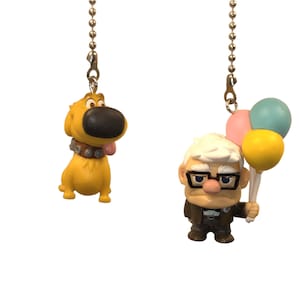 May include: Two ceiling fan pull chains featuring characters from the animated film "Up." One chain has a yellow dog with a brown nose and collar. The other has an older man with glasses holding colourful balloons.