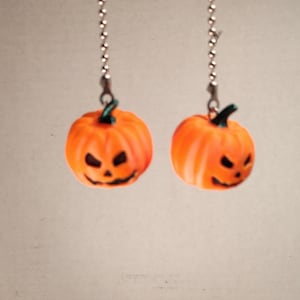May include: A pair of dangling earrings featuring orange jack-o'-lantern pumpkin charms with dark green stems. Each pumpkin has a carved face with triangular eyes and a jagged mouth, suspended from a silver-toned metal ball chain connector.