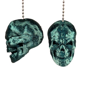 May include: Two teal-colored skull-shaped ceiling fan pulls. One skull is viewed from the side, the other from the front. Each skull is suspended by a silver beaded chain. The skulls have a textured, velvet-like appearance.
