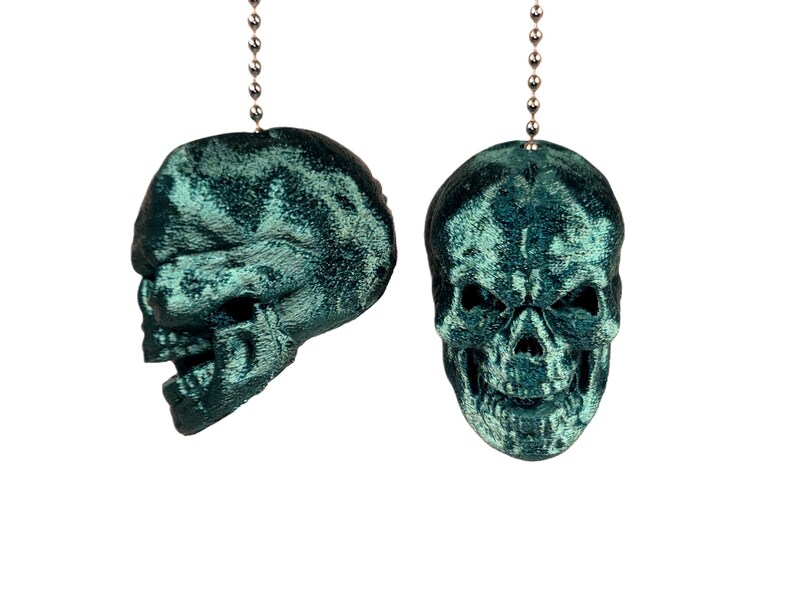 May include: Two teal-colored skull-shaped ceiling fan pulls. One skull is viewed from the side, the other from the front. Each skull is suspended by a silver beaded chain. The skulls have a textured, velvet-like appearance.