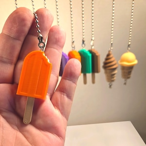 May include: A collection of miniature food-themed keychains hanging from silver ball chains. The foreground features a bright orange popsicle charm with a bite mark, while the background displays various other colorful ice cream and popsicle designs.