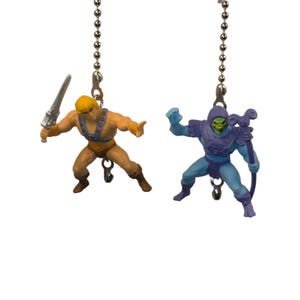 May include: Two miniature action figures of He-Man and Skeletor suspended from silver chains. He-Man holds a sword, Skeletor a staff. Both figures are in dynamic poses, with detailed paintwork.