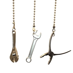 Pode incluir: Três pingentes de ferramentas prateadas pendurados em correntes de bolinhas. As ferramentas incluem uma chave inglesa ajustável, uma chave fixa e um alicate. Fundo branco.