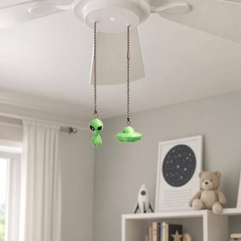 K&ouml;nnte beinhalten: Zwei Deckenventilator-Zugketten mit hellgr&uuml;nen dekorativen Ornamenten. Eine Zugkette endet in einer kleinen Alien-Figur mit gro&szlig;en schwarzen Augen, w&auml;hrend die andere Zugkette in einem gr&uuml;nen, fliegenden Untertassen-Ornament endet.