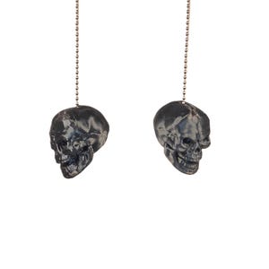 May include: Two black skull-shaped light pull chains hanging against a white background. The skulls have detailed features and are suspended by silver ball chains. The overall aesthetic is gothic and decorative.