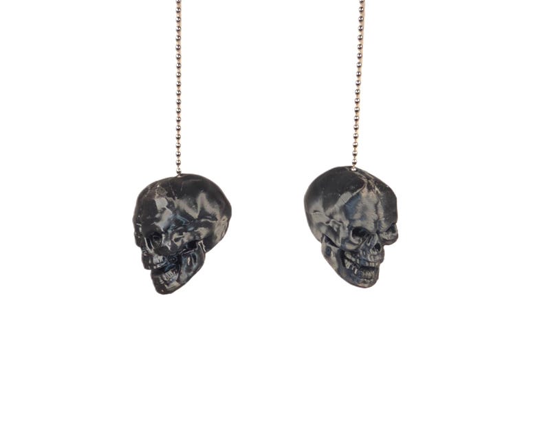 May include: Two black skull-shaped light pull chains hanging against a white background. The skulls have detailed features and are suspended by silver ball chains. The overall aesthetic is gothic and decorative.